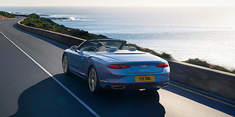 Bentley Jakarta New Bentley Continental GTC Azure convertible rear view in Light Windsor blue paint with 22 inch ten spoke wheels driving dynamically on an ocean road in the sunshine